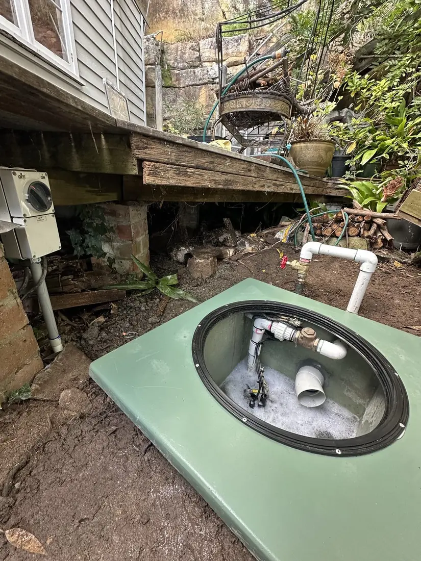 A green fibreglass sewer pump pit at ground level with the man-cover open showing the non-return valve, union and inlet inside, an above-ground gate valve on the discharge, and an electrical isolation switch mounted on the timber beam clear of the pit