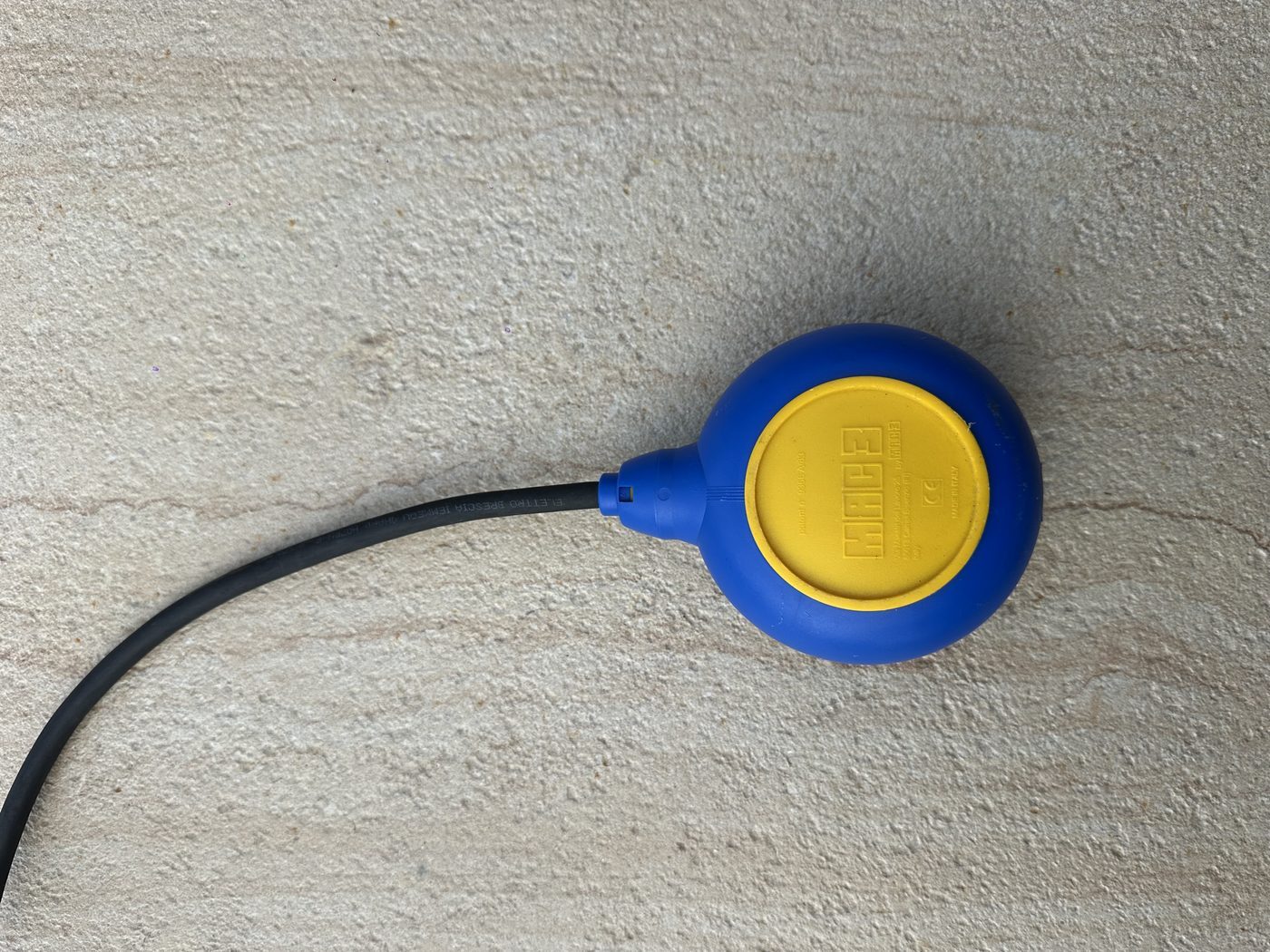 A Mac3 float switch with a blue and yellow moulded body and a heavy black rubber-sheathed cable, resting on a concrete surface