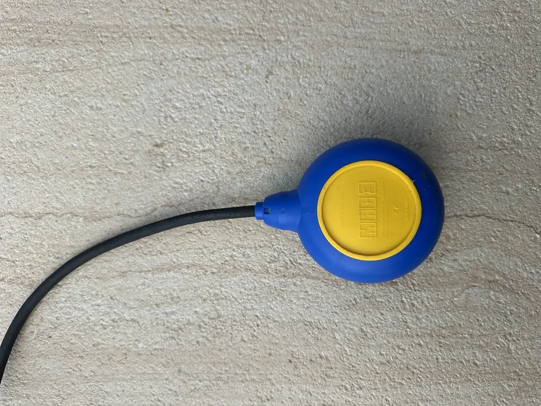 A Mac3 float switch with a blue and yellow moulded body and a heavy black rubber-sheathed cable, resting on a concrete surface