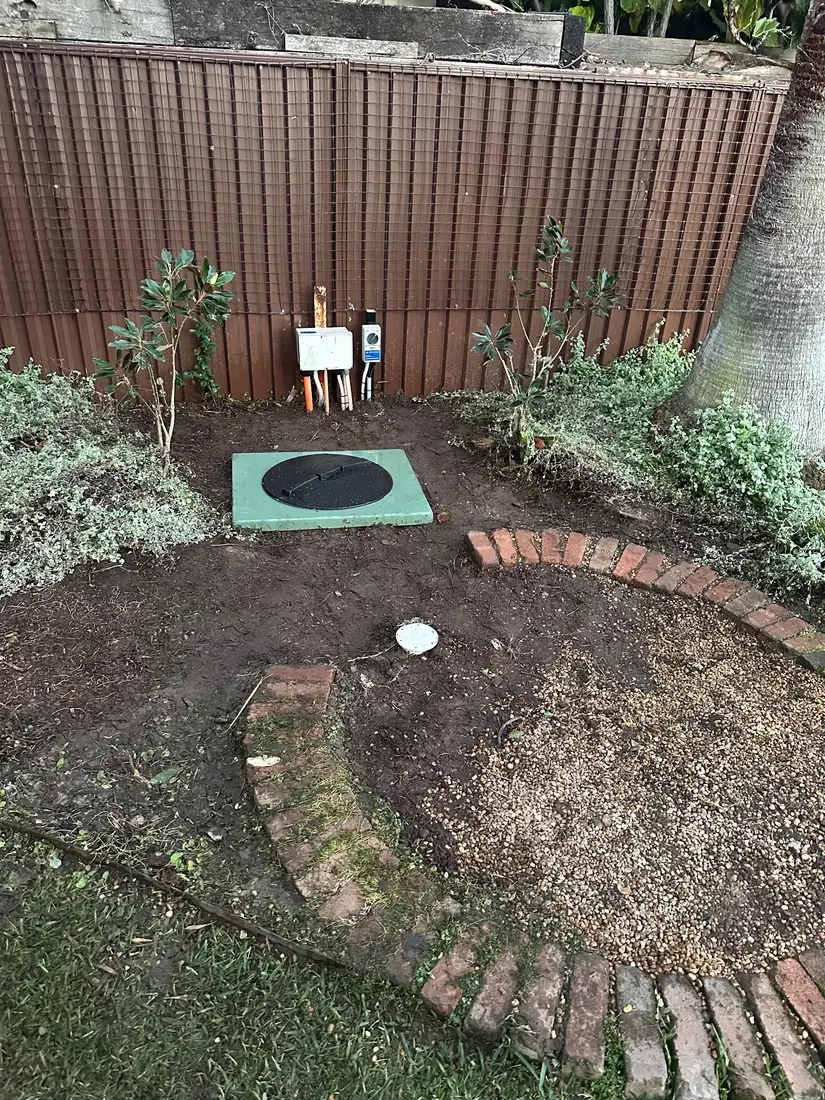 The relocated Burraneer pump pit with new lid and switchgear by the fence, with the old pit's spot backfilled in the garden bed in front