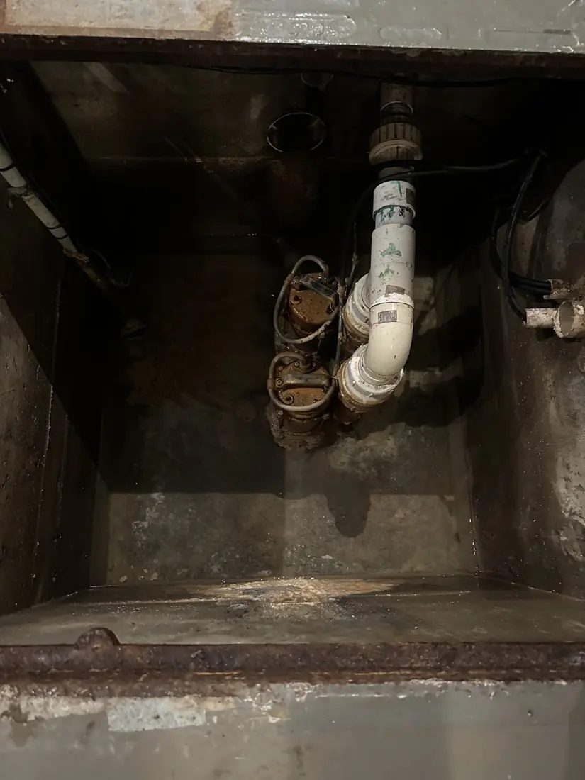 Two old corroded submersible pumps sitting low in a dark pit, the discharge turning up through aging white PVC pipework
